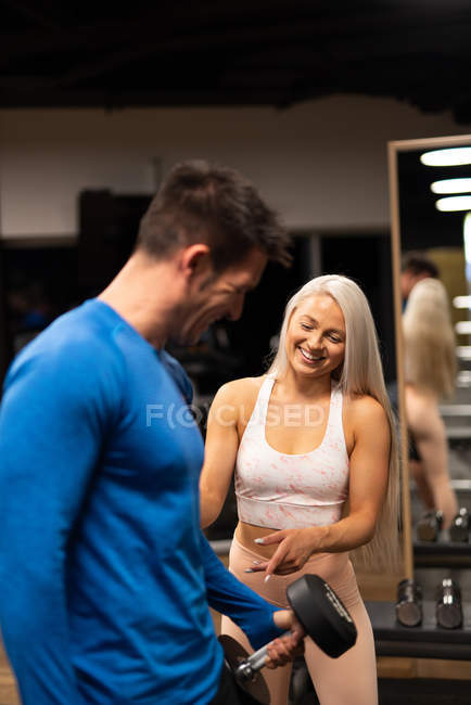 Mulher ajudando o homem a fazer exercício com halteres no ginásio — Fotografia de Stock