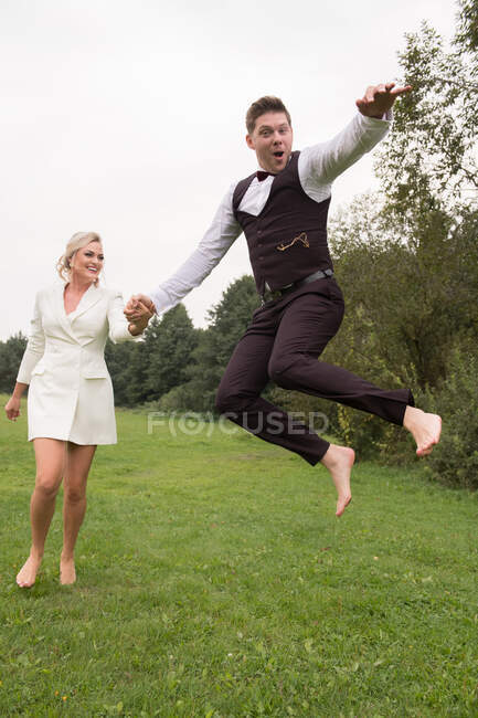 Trendy adult groom and bride in elegant suits holding hands and jumping with excitement on green meadow — Stock Photo