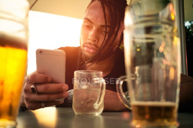 Crop view of young African-American guy holding and looking at mobile phone, sitting in cafe near glasses with beer in Austria — Stock Photo
