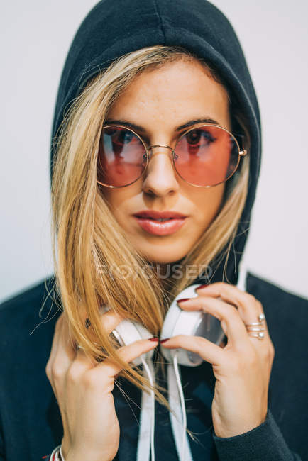 Giovane donna bionda con cappuccio nero e occhiali da sole rotondi con cuffie sul collo guardando la fotocamera su sfondo bianco — Foto stock