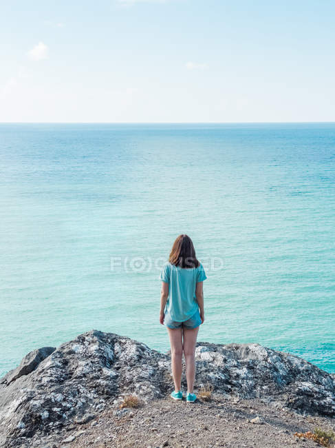 Mulher em shorts de pé na borda do penhasco acima da água do mar azul sem fim — Fotografia de Stock
