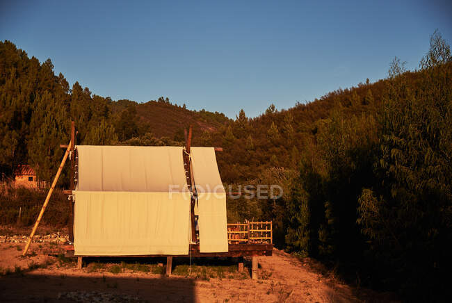 Tienda de tela beige en plataforma de madera con valla de pie en el prado en el bosque espeso resaltado por la puesta del sol con el cielo azul en el fondo - foto de stock