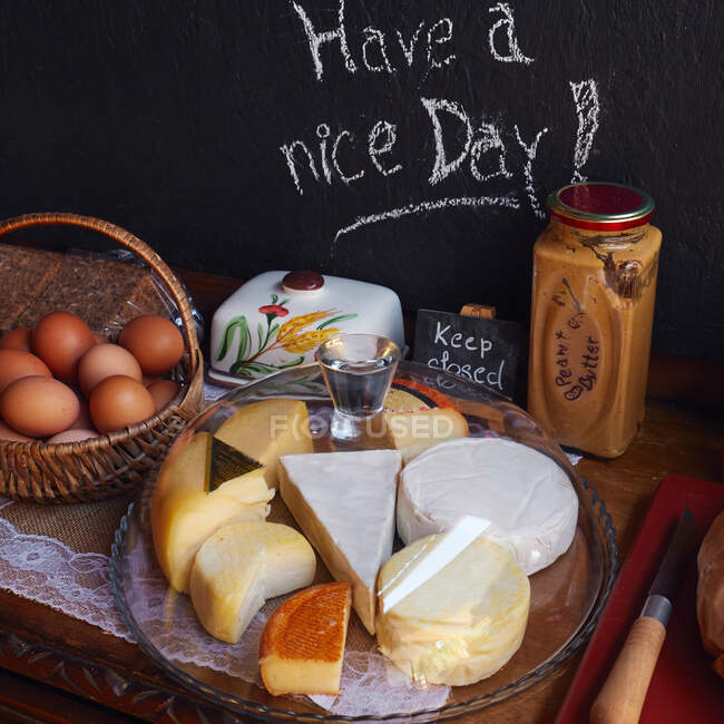 Plate with many kinds of cheese and basket with eggs with peanut butter standing on wooden table with chalkboard on background — Stock Photo