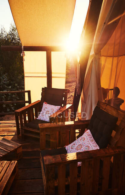 Cadeiras de madeira marrom com travesseiro colorido no chão de madeira com tenda bege? e folhagem verde com raios de sol no fundo — Fotografia de Stock