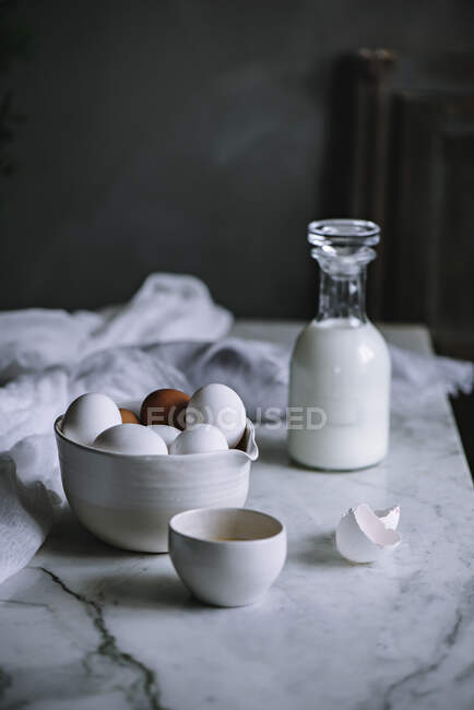 Bol d'œufs de poulet et bouteille de produits laitiers frais debout sur une table en marbre dans la cuisine — Photo de stock