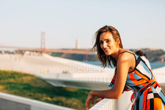 Femme élégante souriante en robe appuyée sur la clôture du pont et observant le paysage urbain à la lumière du soleil — Photo de stock