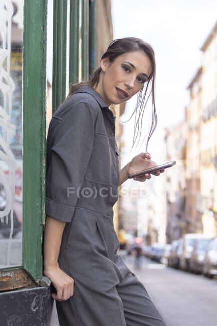 Vista laterale di bella femmina in abito elegante navigando smartphone moderno e guardando la fotocamera mentre si appoggia sul palazzo sulla strada della città — Foto stock