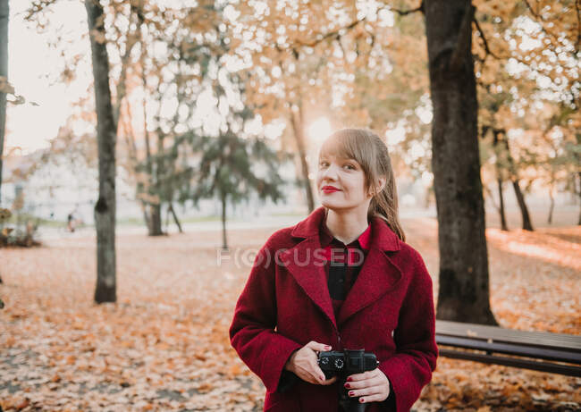 Jeune femme en manteau rouge tenant appareil numérique et regardant la caméra dans la forêt d'automne — Photo de stock