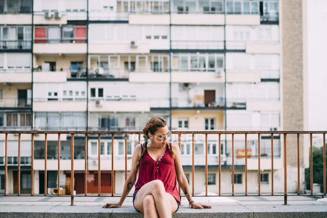 Élégante fille aux cheveux roux avec des tresses et des lunettes de soleil assis près de balustrade contre le bâtiment résidentiel — Photo de stock