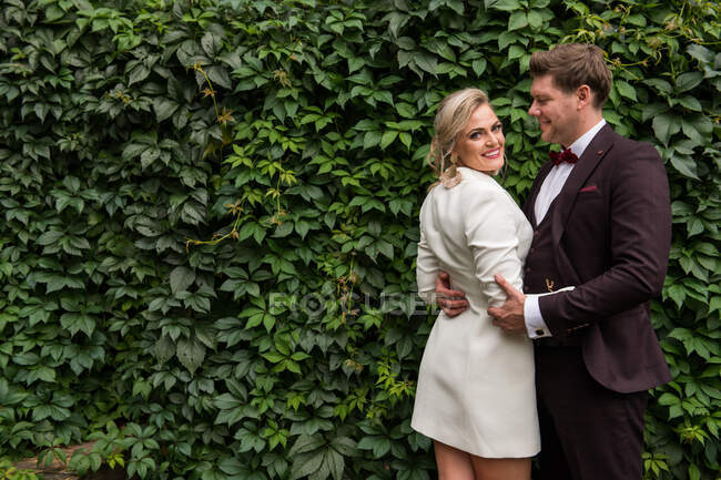 Elegante erwachsene Braut und Bräutigam umarmen sich, während sie in Anzügen vor einer Holzwand stehen und in die Kamera lächeln — Stockfoto