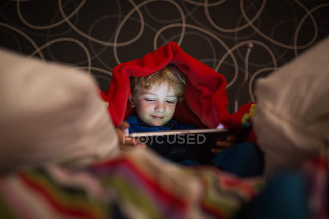 Smiling little boy in pajamas using digital tablet under blanket — Stock Photo