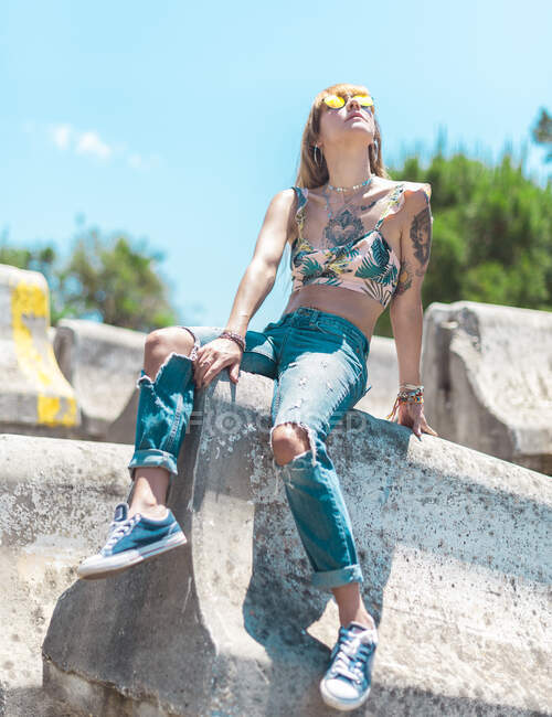 Fit tattooed female in yellow sunglasses, torn jeans and sneakers sitting on concrete plates and looking up — Stock Photo