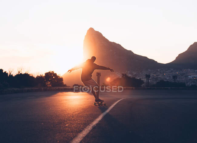 Vista posteriore dell'uomo che cavalca sullo skateboard su strada asfaltata giù per la collina in controluce. — Foto stock