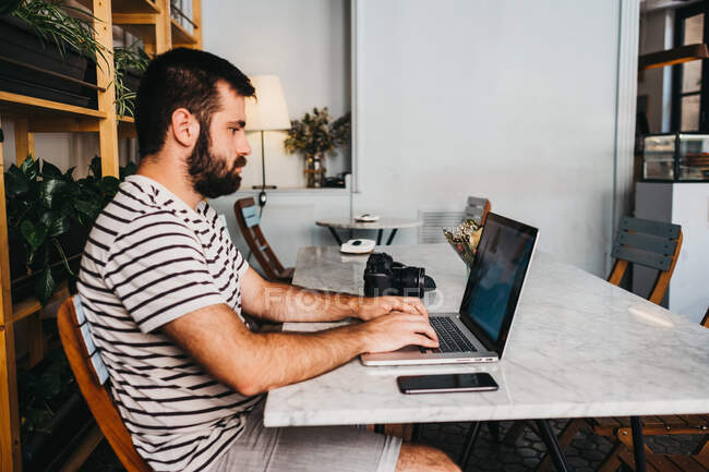 Вид сбоку на бородатого мужчину с фотокамерой и смартфоном, сидящего за столом при использовании ноутбука — стоковое фото