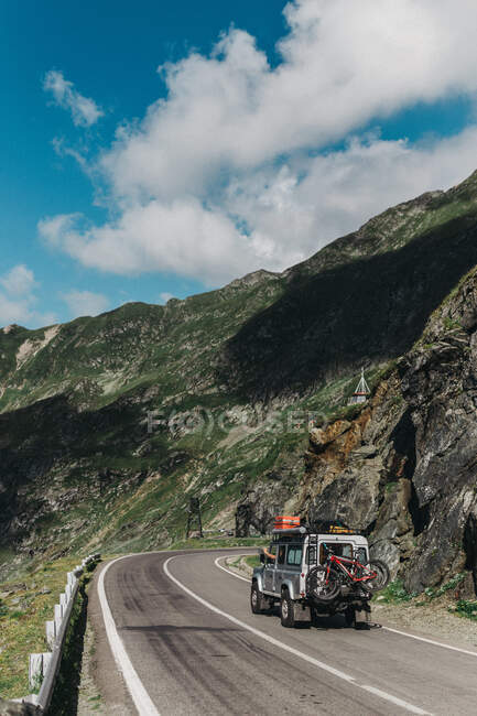 Off-road vehicle with bicycle riding narrow asphalt road near mountains on sunny day — Stock Photo