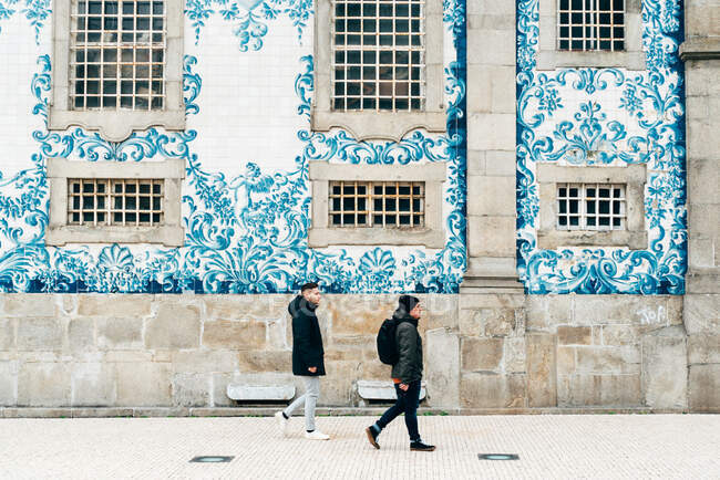 Vue latérale de touristes joyeux marchant au mur décoré de carreaux blancs et bleus. — Photo de stock