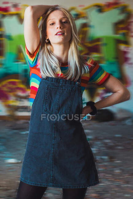 Young trendy blond model standing in abandoned building with walls in graffiti looking confidently at camera. — Stock Photo