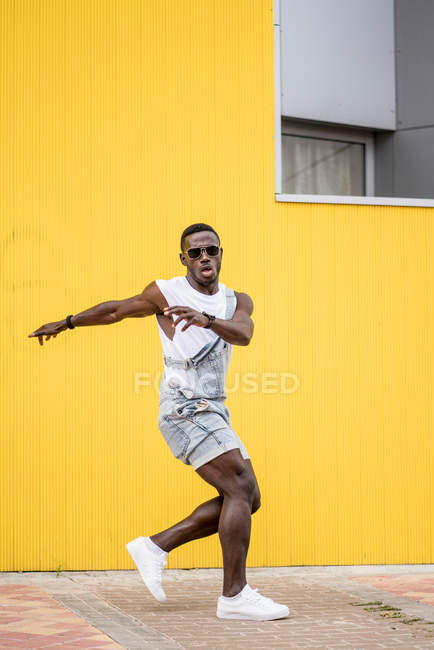 Hombre afroamericano en traje de mezclilla practicando break dance sobre fondo amarillo - foto de stock