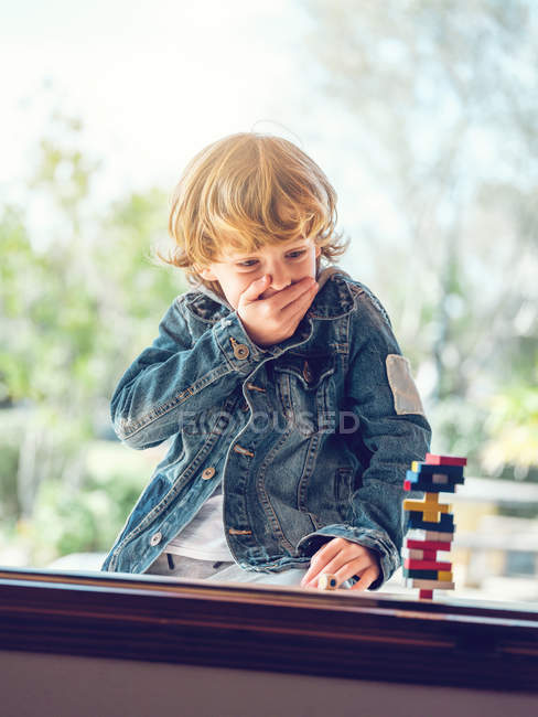 Bonito menino surpreso sentado na janela e brincando com blocos de torre de madeira — Fotografia de Stock