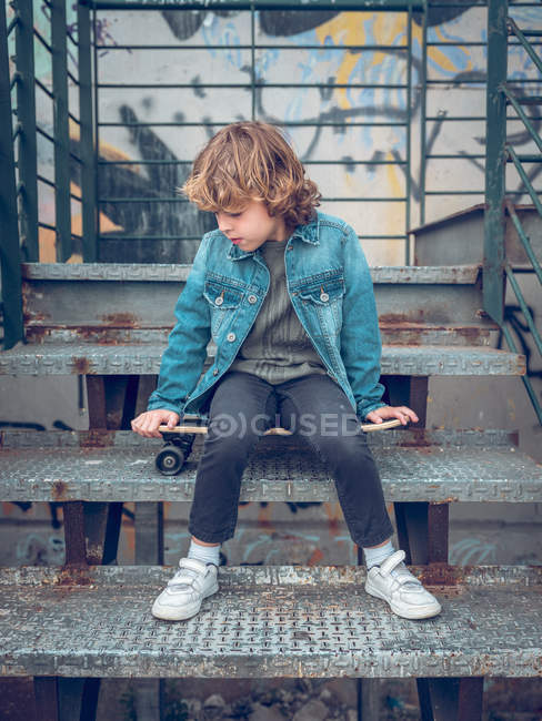 Garçon réfléchi assis sur les escaliers avec planche à roulettes — Photo de stock