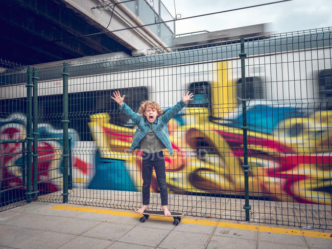 Funny barefoot boy on skateboard with rising hands in front of train — Stock Photo