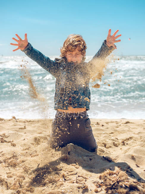 Молодий хлопчик грає і кидає пісок на тлі океану в сонячний день — стокове фото