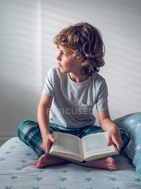 Симпатичний хлопчик у піжамі сидить на зручному ліжку і читає гарну книгу — стокове фото
