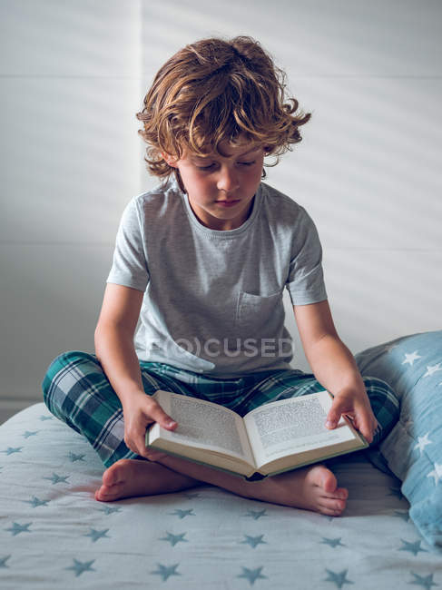 Симпатичный мальчик в пижаме сидит на удобной кровати и читает хорошую книгу — стоковое фото