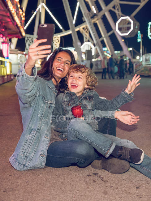 Fröhliche Mutter und kleiner Sohn sitzen auf dem Boden und machen Selfie auf Jahrmarkt — Stockfoto