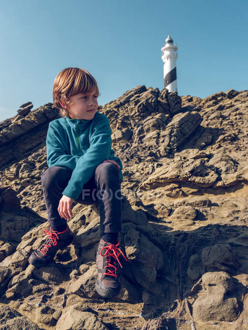 Young boy sitting on rocky hill on background of beacon tower — Stock Photo