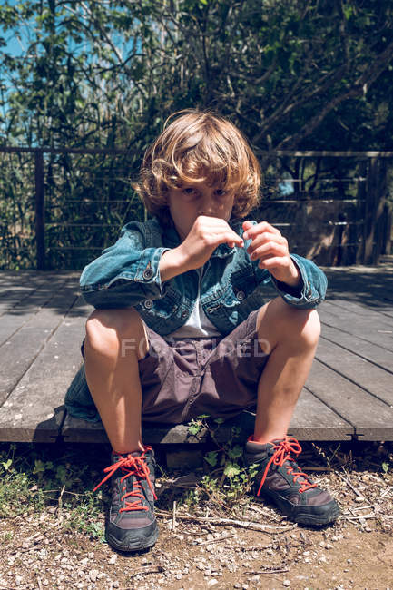 Pensive little boy with curly blond hair sitting on wooden footpath in countryside — Stock Photo