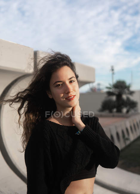 Adorável jovem mulher com cabelo encaracolado sorrindo e tocando o pescoço enquanto estava de pé no fundo borrado da rua da cidade — Fotografia de Stock