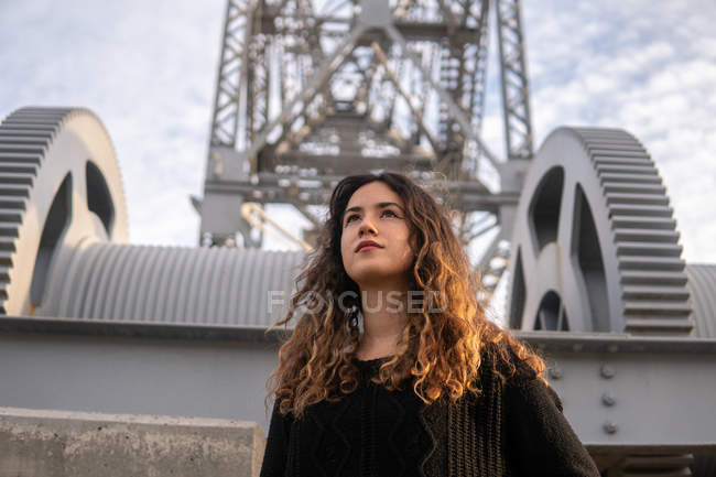 Attraktive junge Frau mit lockigem Haar, die wegschaut, während sie auf dem Hintergrund auf moderner Struktur und bewölktem Himmel auf der Stadtstraße steht — Stockfoto