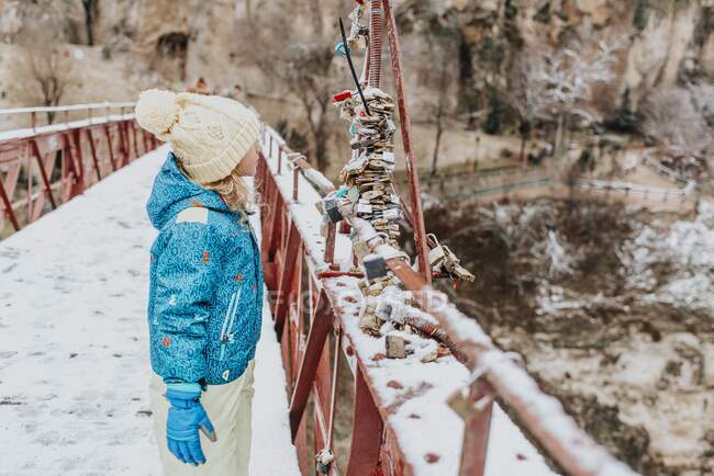 Vista lateral da criança bonita em roupas quentes em pé na ponte moderna e olhando para um monte de cadeados de amor no dia de inverno no campo — Fotografia de Stock