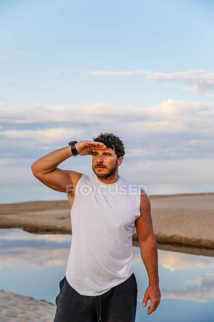 Vollbärtiger erwachsener Mann in Sportbekleidung, der die Hand nahe der Stirn hält und beim Outdoor-Training am Strand in die Ferne schaut — Stockfoto
