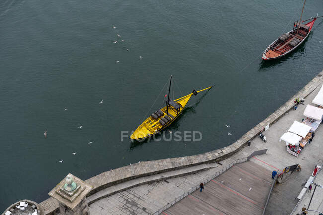 Dall'alto argine della città con persone che camminano, navi che galleggiano sulla superficie dell'acqua e uccelli volanti in Oporto, Portogallo — Foto stock