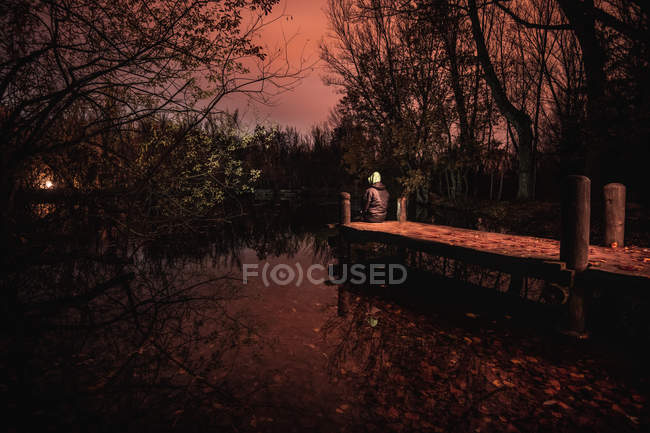 Atrás vista hombre en el capó sentado en el muelle cerca del lago entre el oscuro bosque sombrío con el cielo rojo en Bosque Finlandes, Rascafria, España - foto de stock
