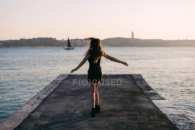 Cheerful woman in black wear and boots dancing on embankment near water surface with yacht at sunset — Stock Photo