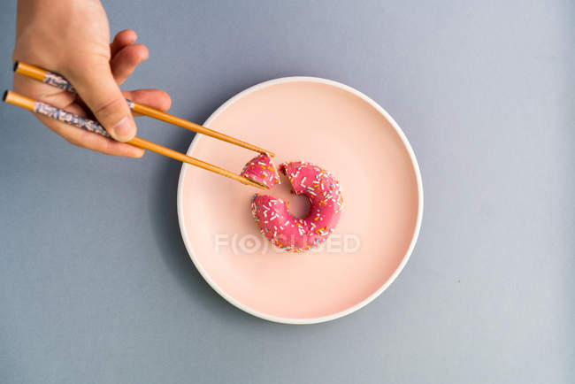 Зверху рука людини з паличками, що тримають скибочку смачного пончика на блюді на синьому фоні — стокове фото