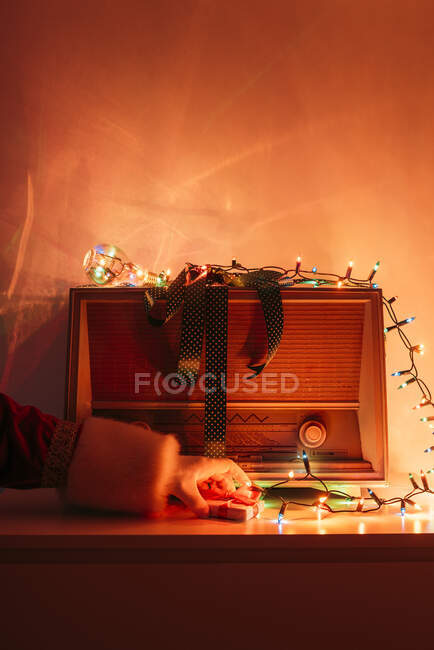 Hand of anonymous Santa Claus putting small present on cabinet near vintage radio receiver and colorful lights — Stock Photo