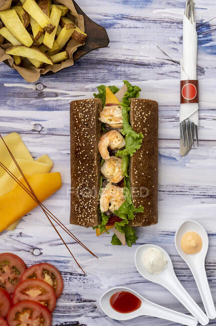 From above view of sliced vegetables and cheese with big sandwich placed in middle on wooden background — Stock Photo