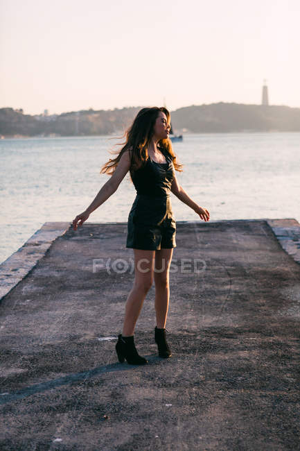 Dreamy charming young woman in black wear and boots dancing on embankment near water surface at sunset — Stock Photo