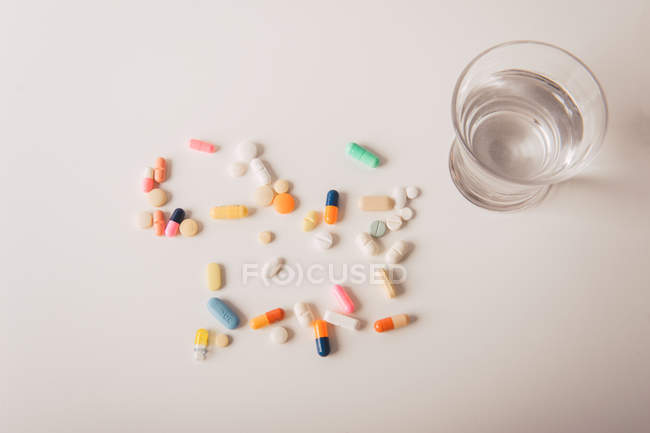 Різнокольорові таблетки та капсули, розкидані склянкою води на білому тлі — стокове фото
