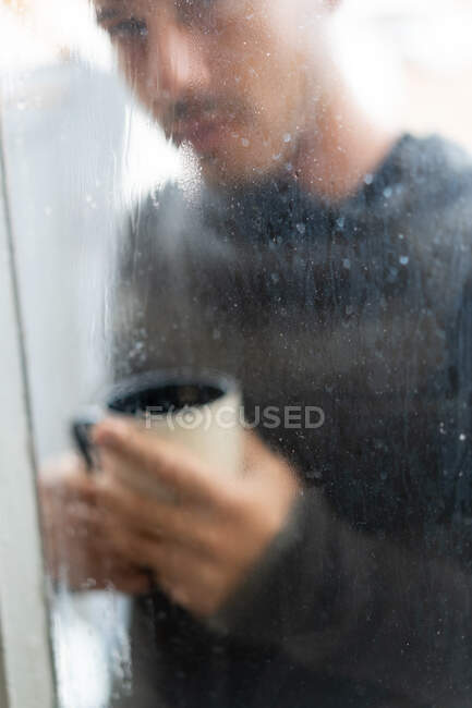 Tiro através de vidro janela molhada do homem em pé suéter com xícara de café — Fotografia de Stock