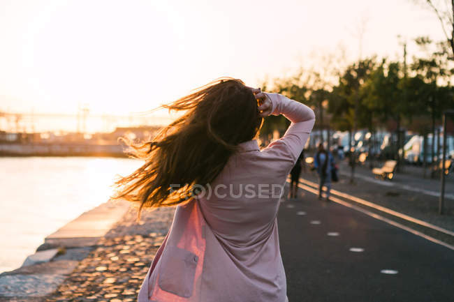 Вид сзади на девушку, идущую по набережной возле воды и переулка на закате — стоковое фото