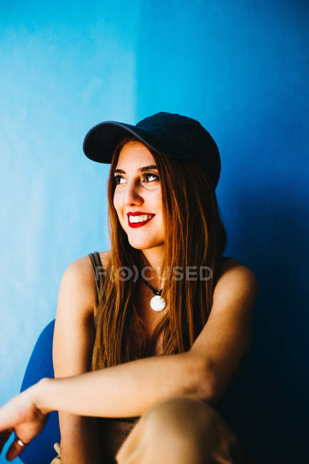 Giovane donna dai capelli rossi allegri in berretto e canotta guardando lontano e seduto vicino al muro blu — Foto stock