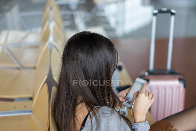 Giovane donna sorridente con auricolari utilizzando smartphone e seduta vicino alla finestra — Foto stock