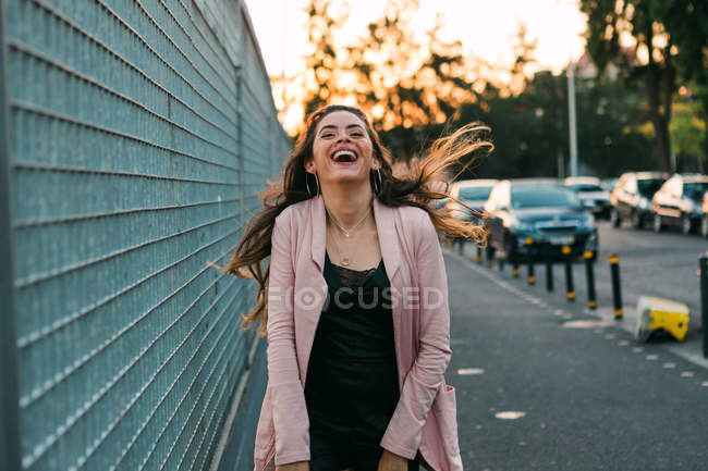 Смеющаяся молодая женщина, идущая по улице возле машин на закате — стоковое фото