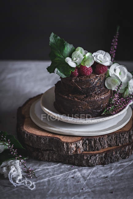 Gâteau au chocolat décoré de framboises et de fleurs servi sur assiette sur support en bois — Photo de stock