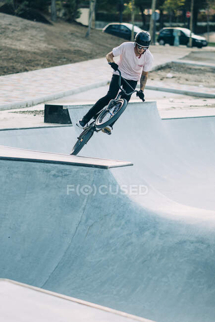 Young man in white T-shirt and helmet riding BMX on springboard on background of road and cars — Stock Photo